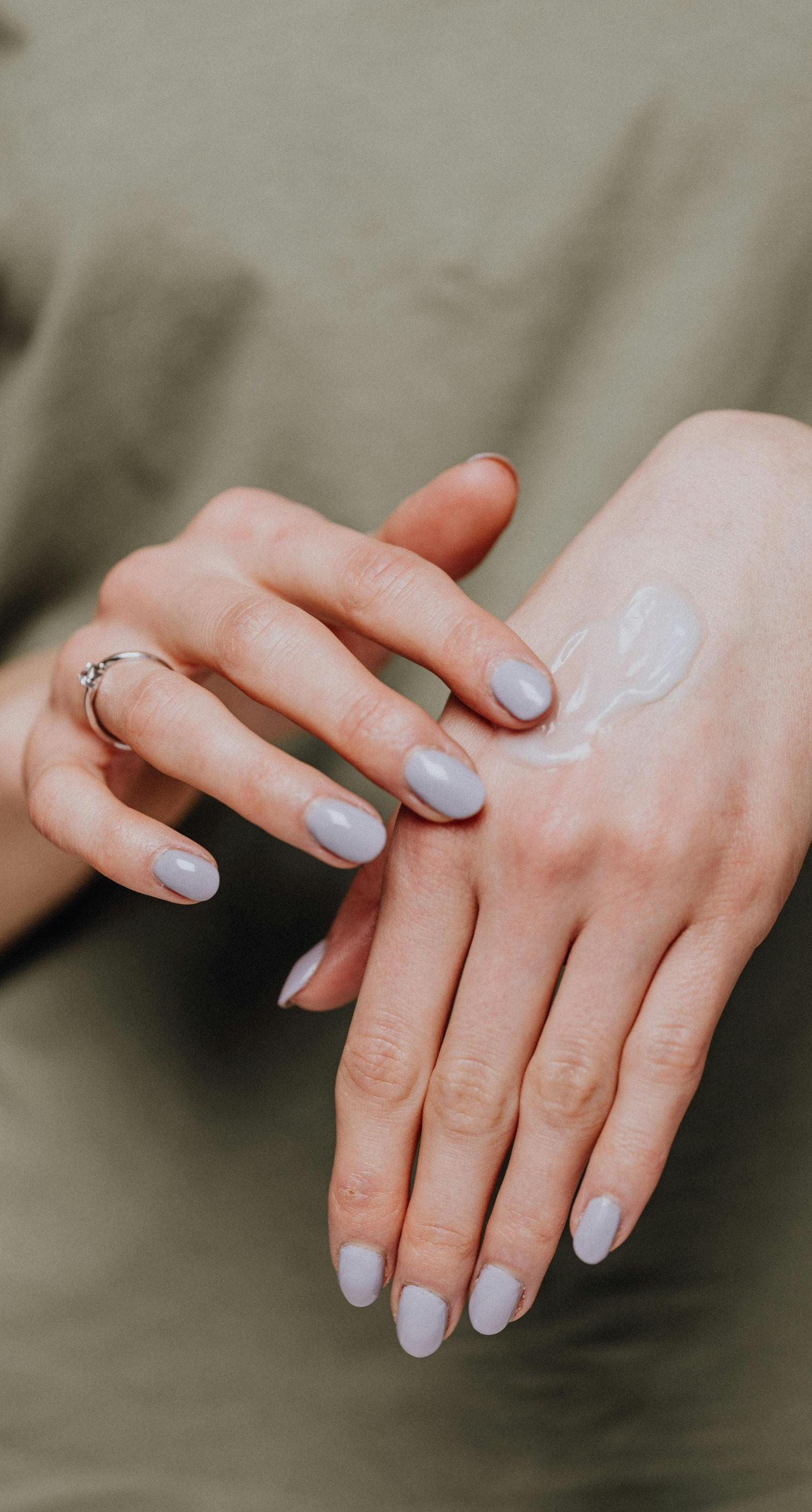 Person applying cream to their hand, close-up. Light skin, gray nails, silver ring, green shirt.