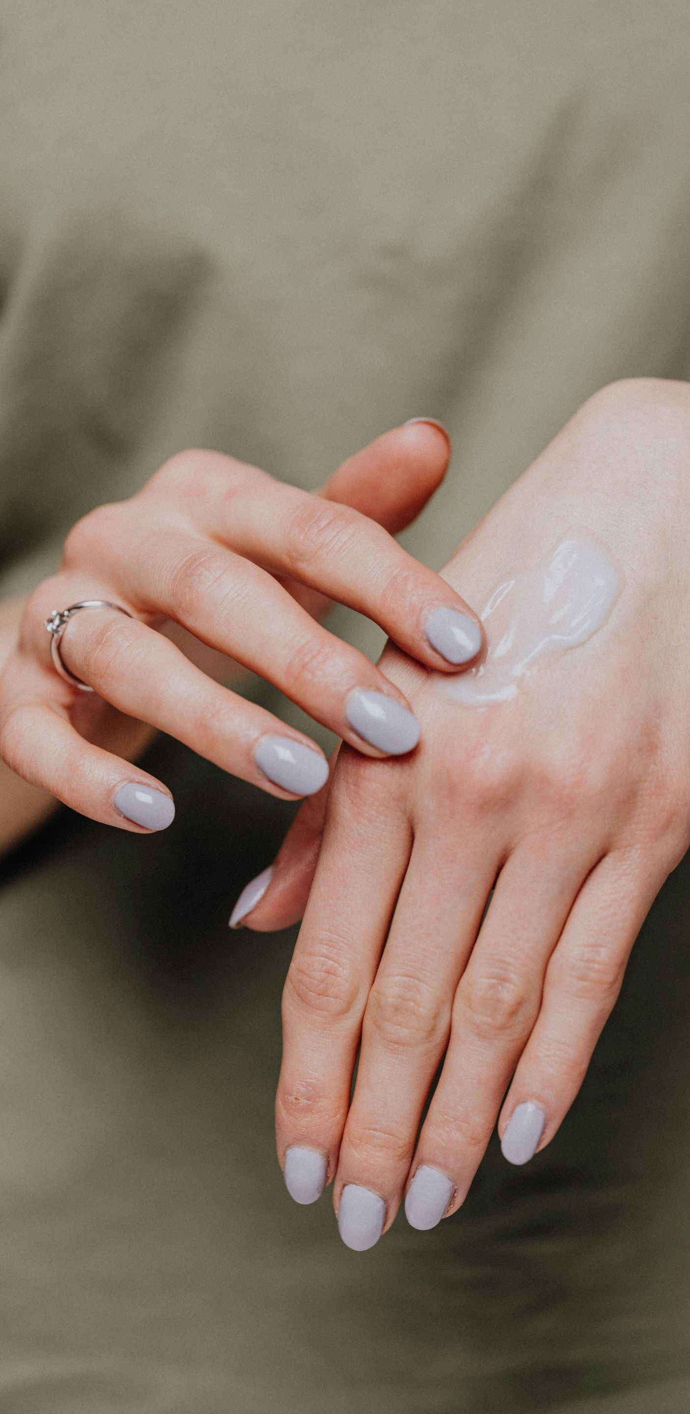 Person applying cream to their hand, close-up. Light skin, painted nails, wearing a ring, against a green shirt.
