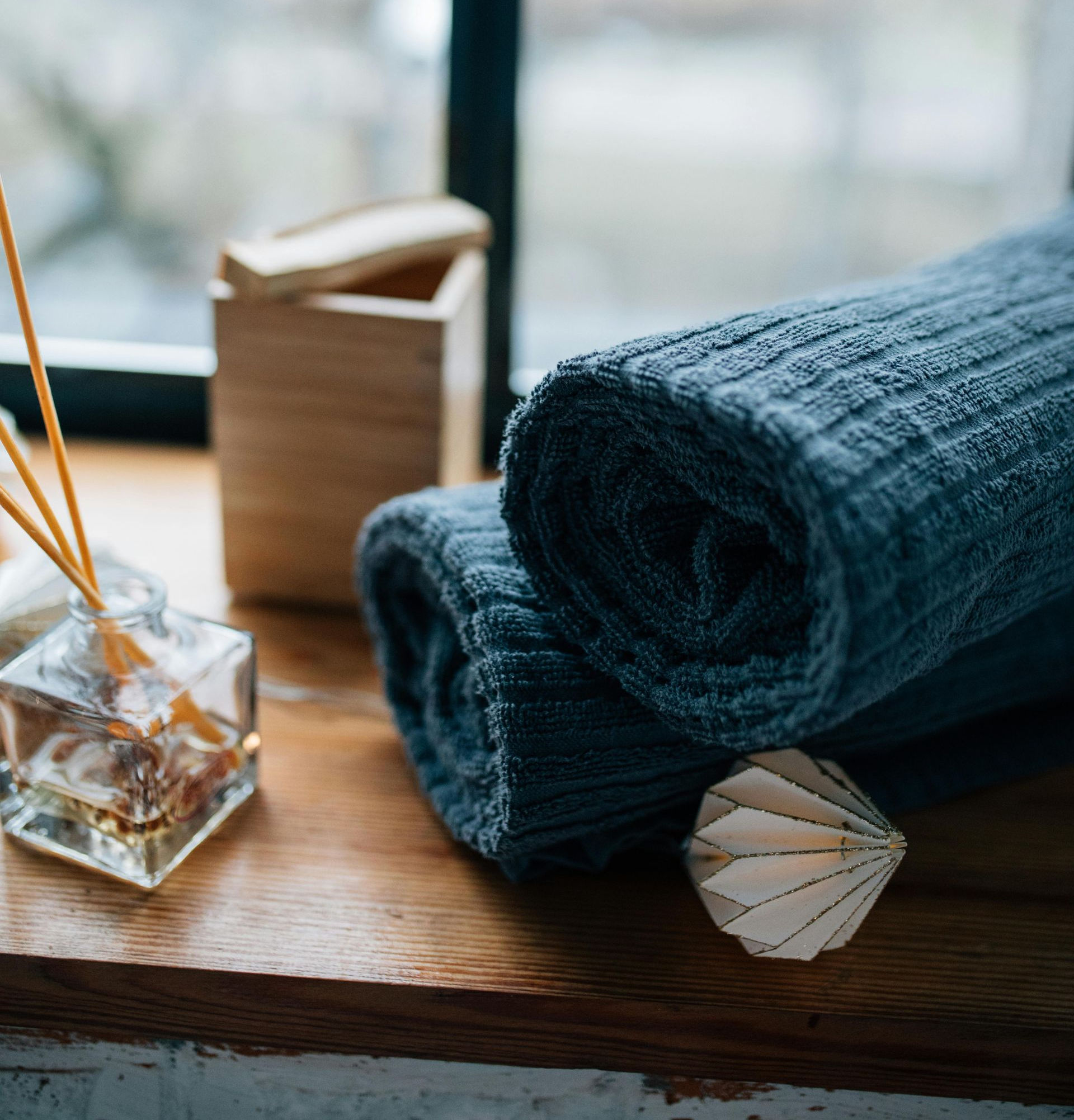 Rolled blue towels on a wooden surface with a diffuser, wooden box, and paper ornament by a window.
