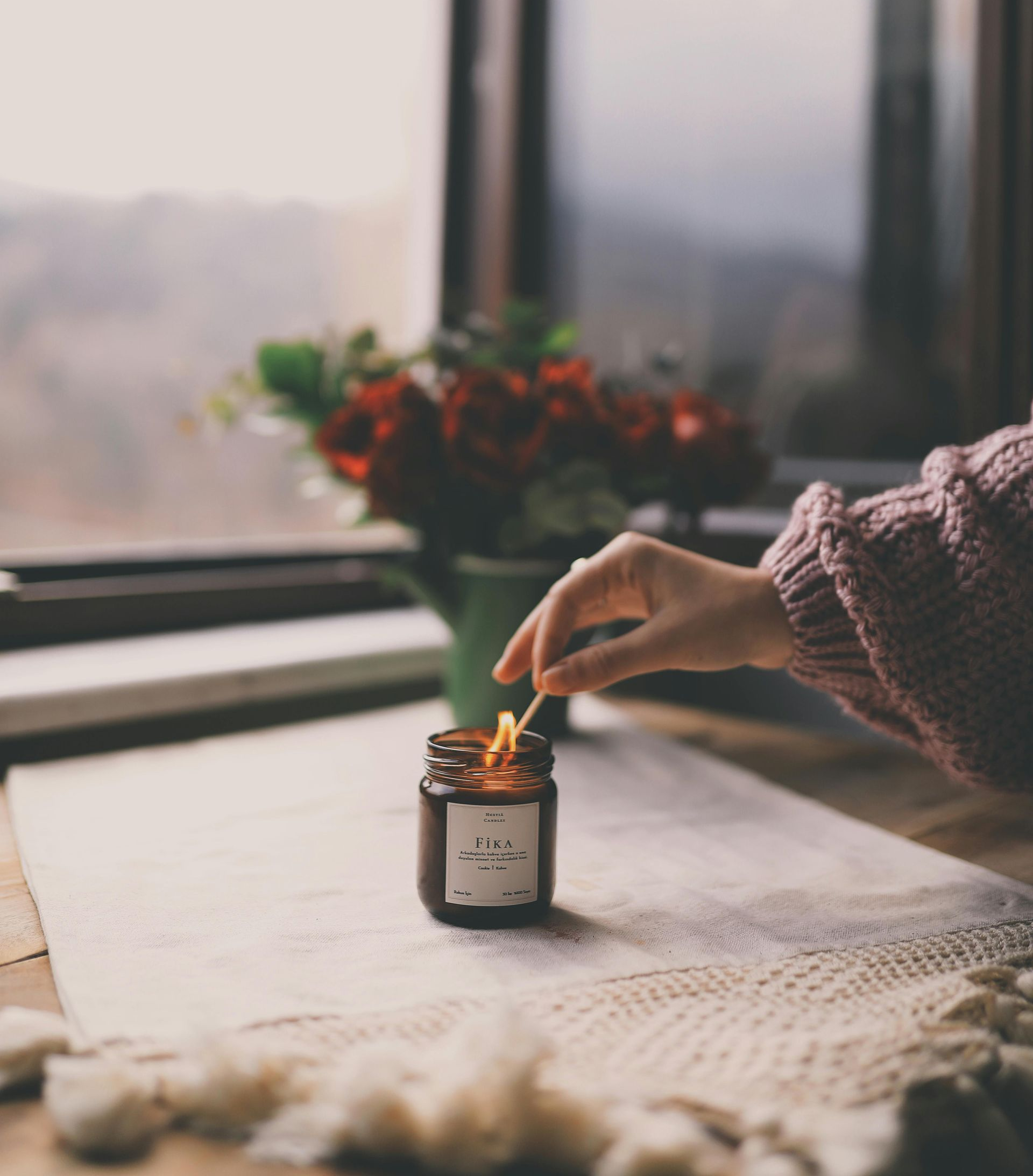 Hand lighting a candle next to a bouquet of flowers by a window. Soft lighting, cozy interior.