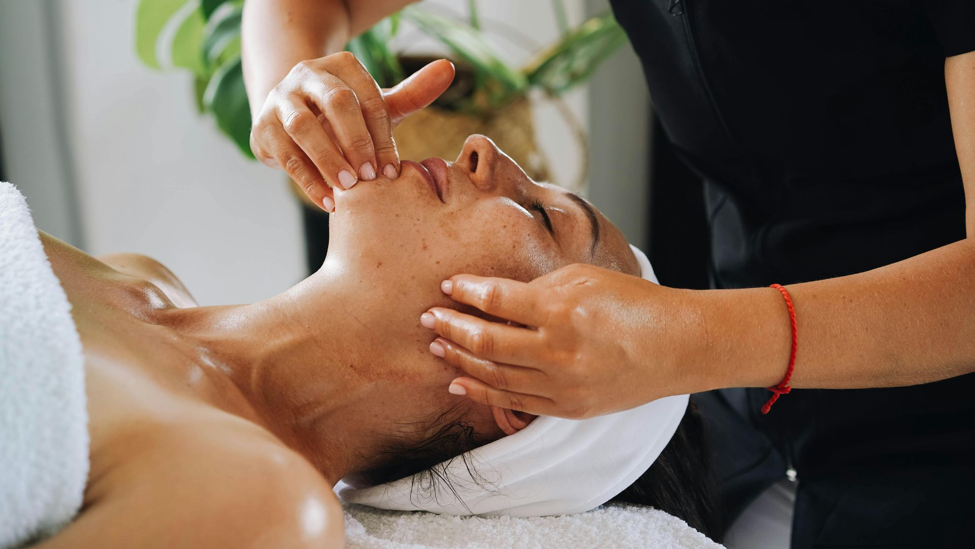 Person receiving facial massage in a spa, hands on face, white towel, plant background.