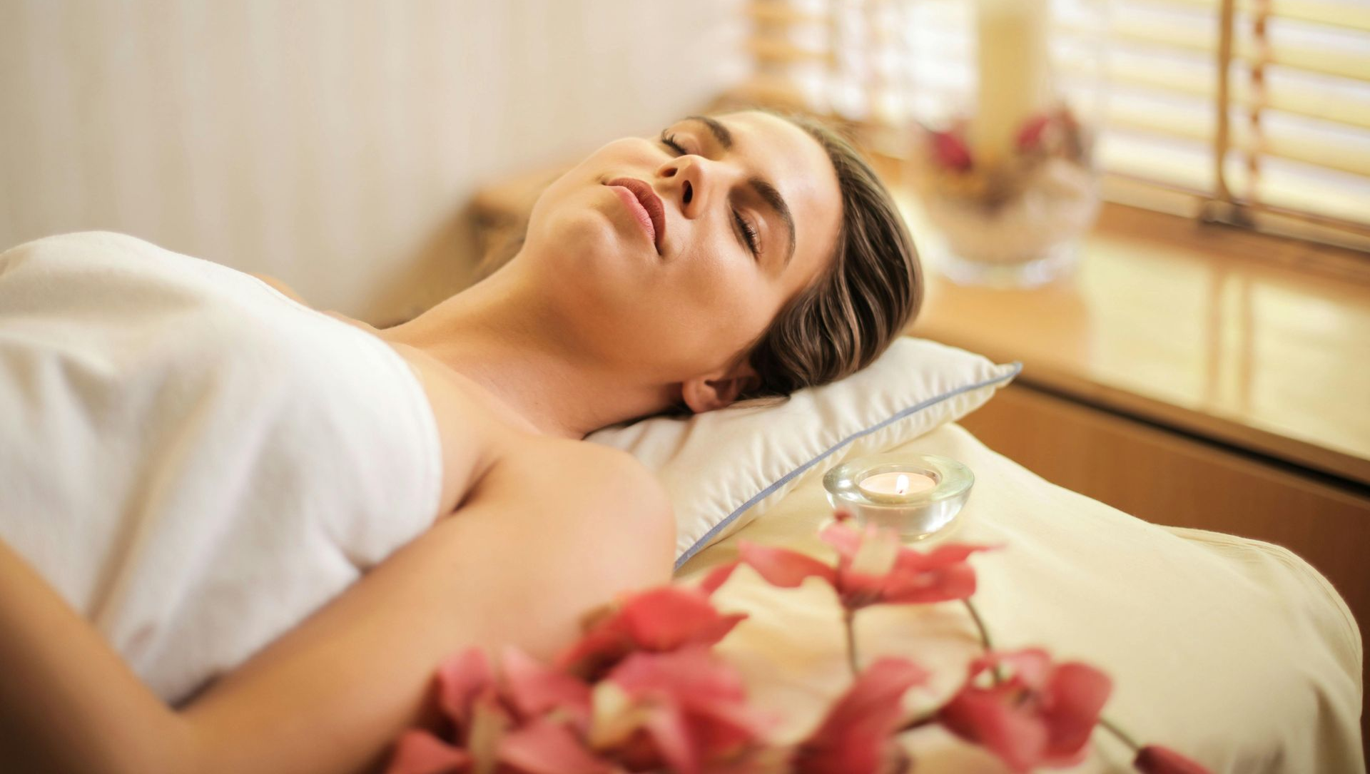 Woman relaxing on massage table, wrapped in white towel, eyes closed, spa setting.