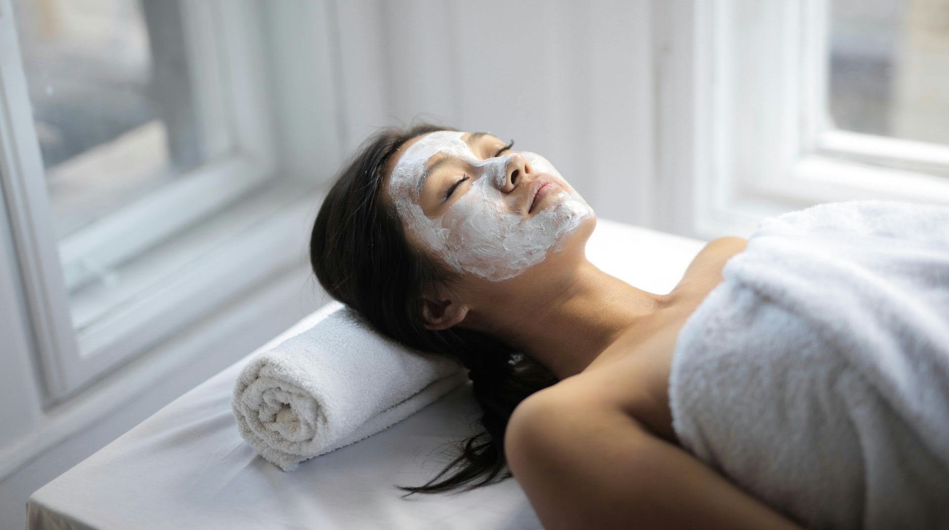 Woman with facial mask relaxing on spa bed near a window.