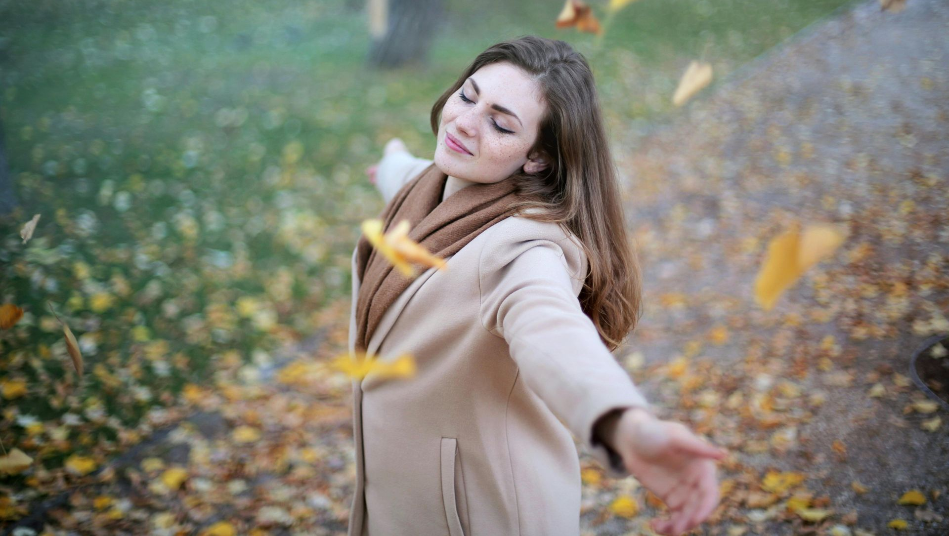 Woman with outstretched arms, eyes closed, surrounded by falling autumn leaves in a park.