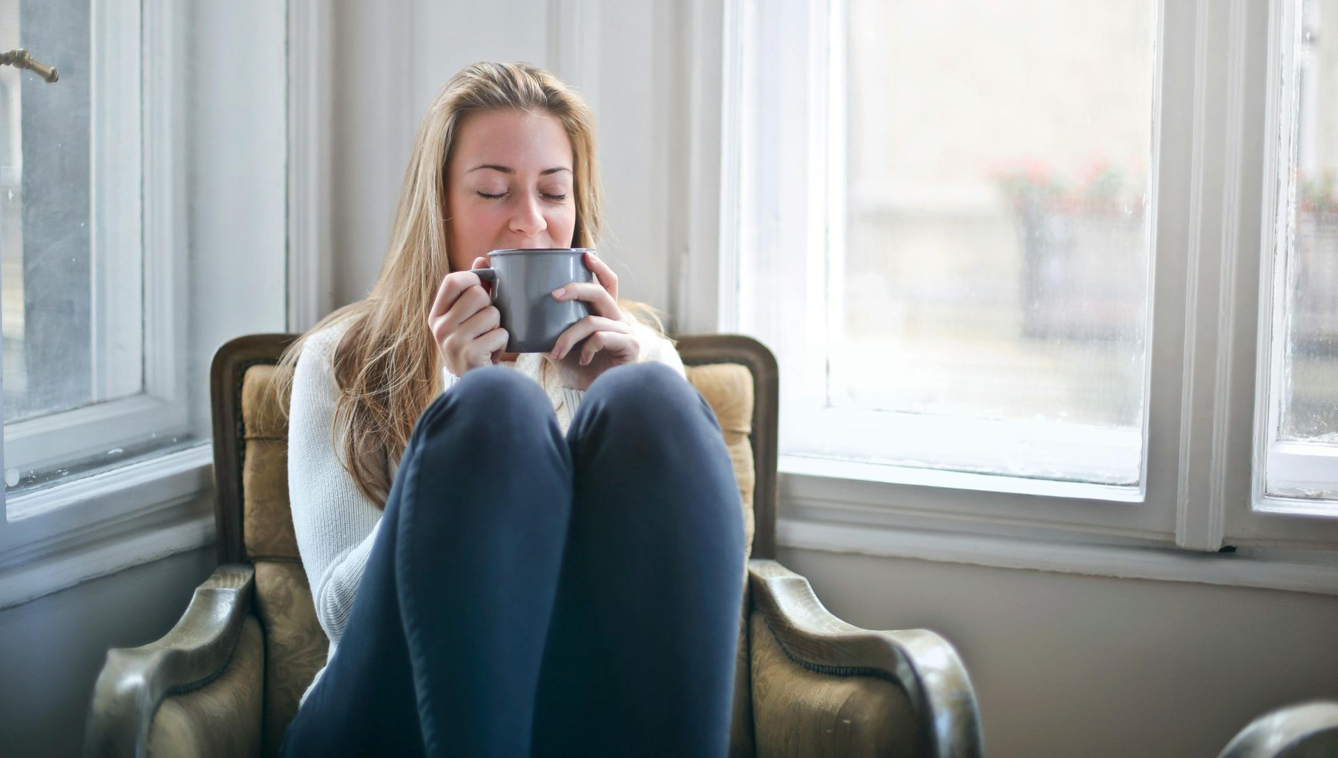 Woman sitting in chair, holding mug near window. Enjoying drink, relaxed expression.