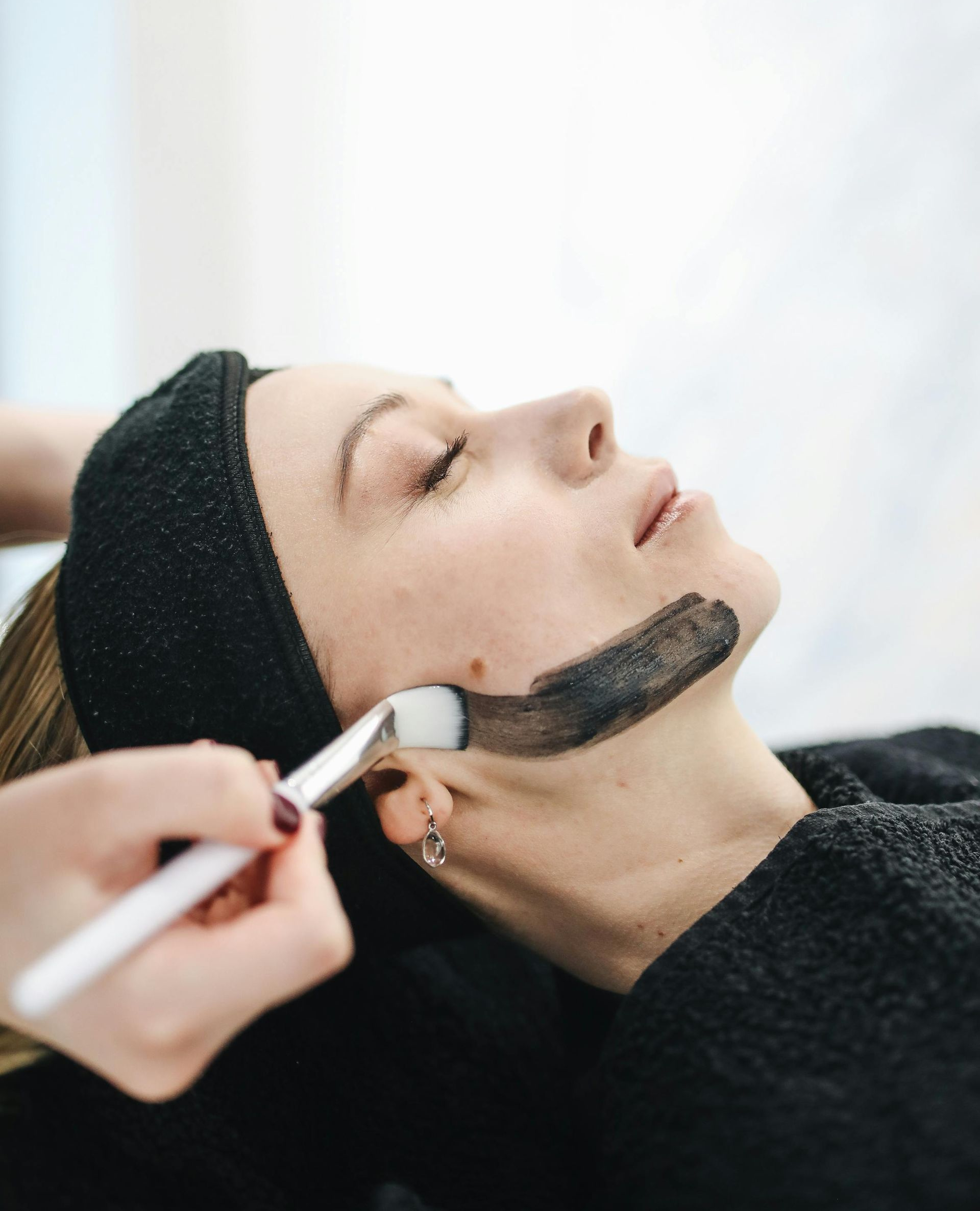 Woman receiving facial treatment, charcoal mask applied with a brush.