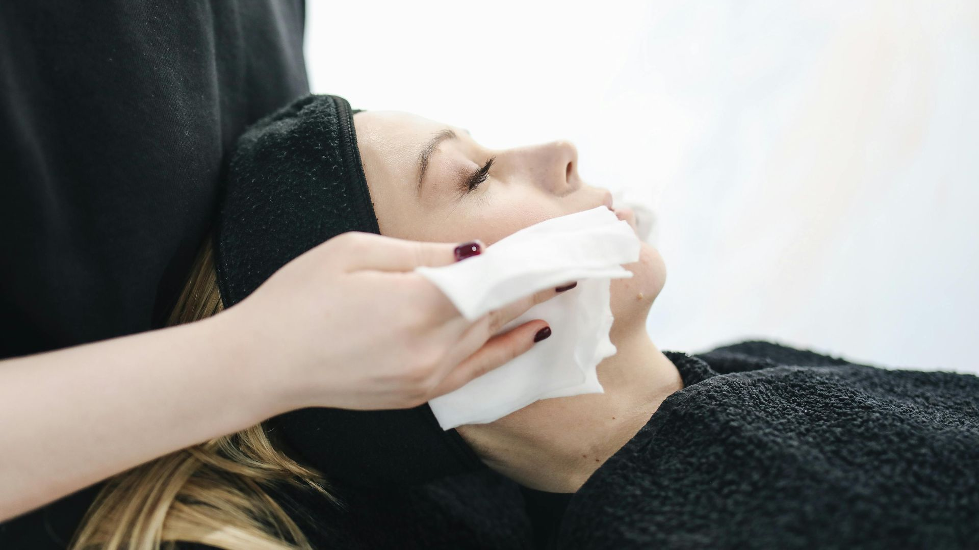 Person receiving facial treatment; wipes being used on the face, indoors, black headband and gown.
