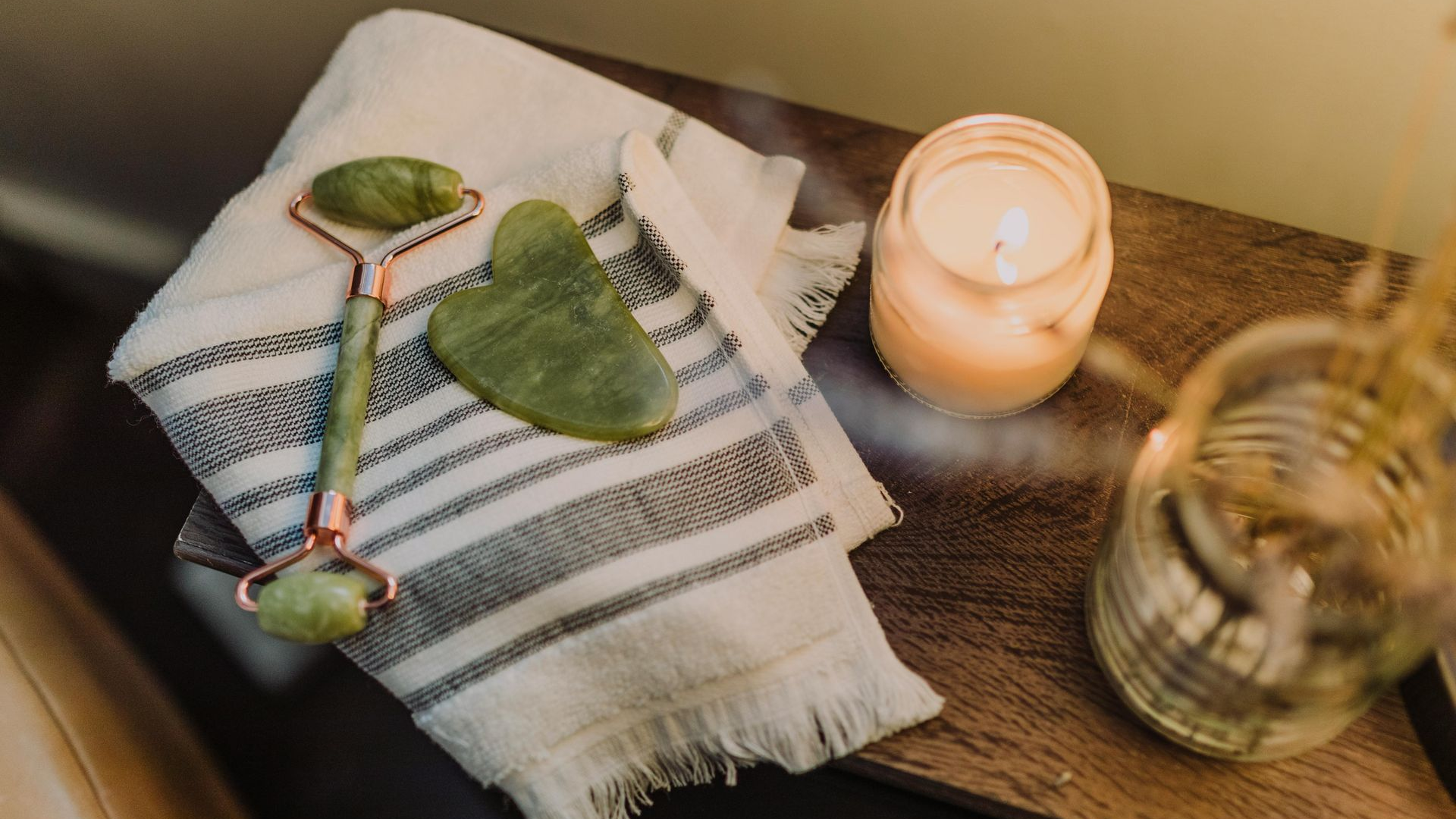Jade facial roller and Gua Sha on towel with candle and glass jar on wooden surface.