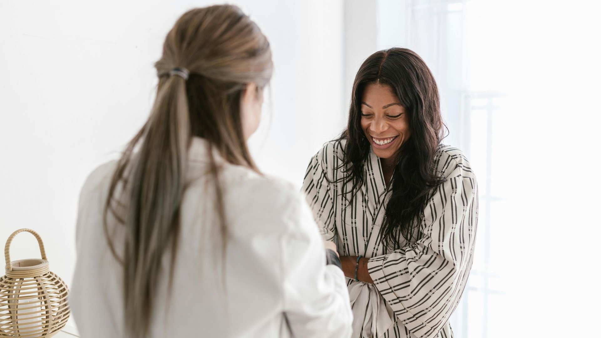 Woman in robe laughs while talking to a person in a white coat, light-filled room.
