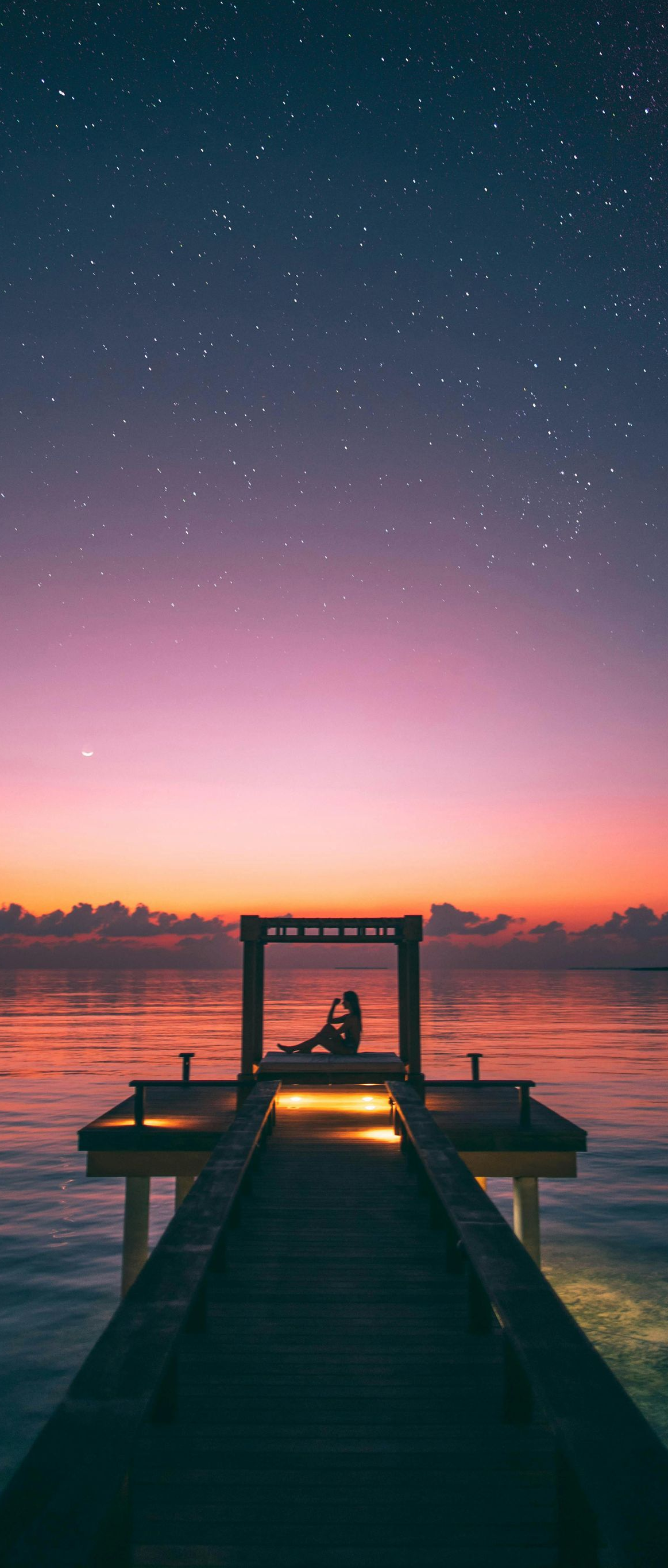 A person sits on a pier, gazing at a starry sky and a colorful sunset over the water.