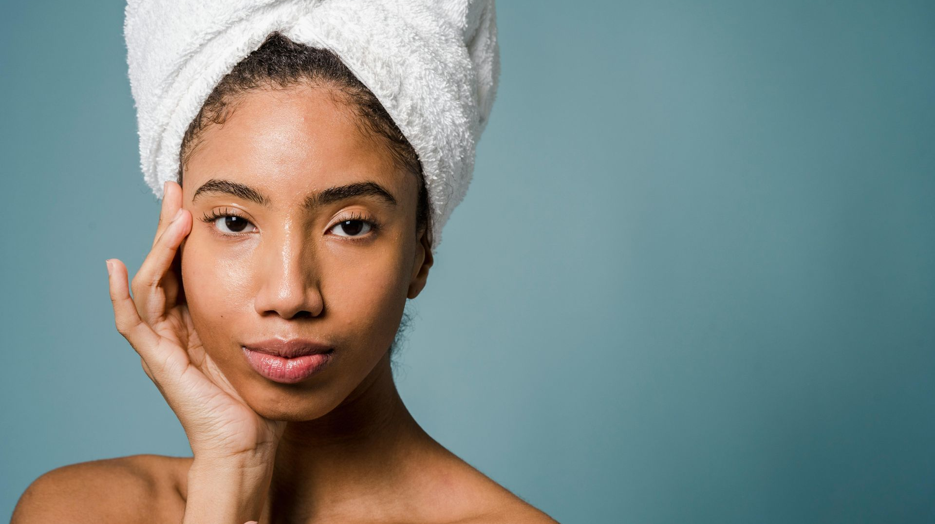 Woman with a white towel on her head, touching her face, against a blue background.