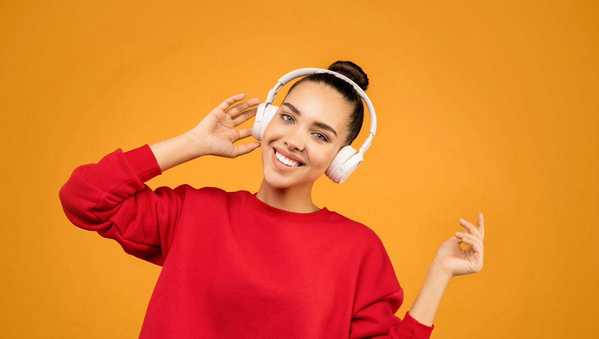 Woman wearing white headphones and red sweatshirt smiles against orange backdrop.