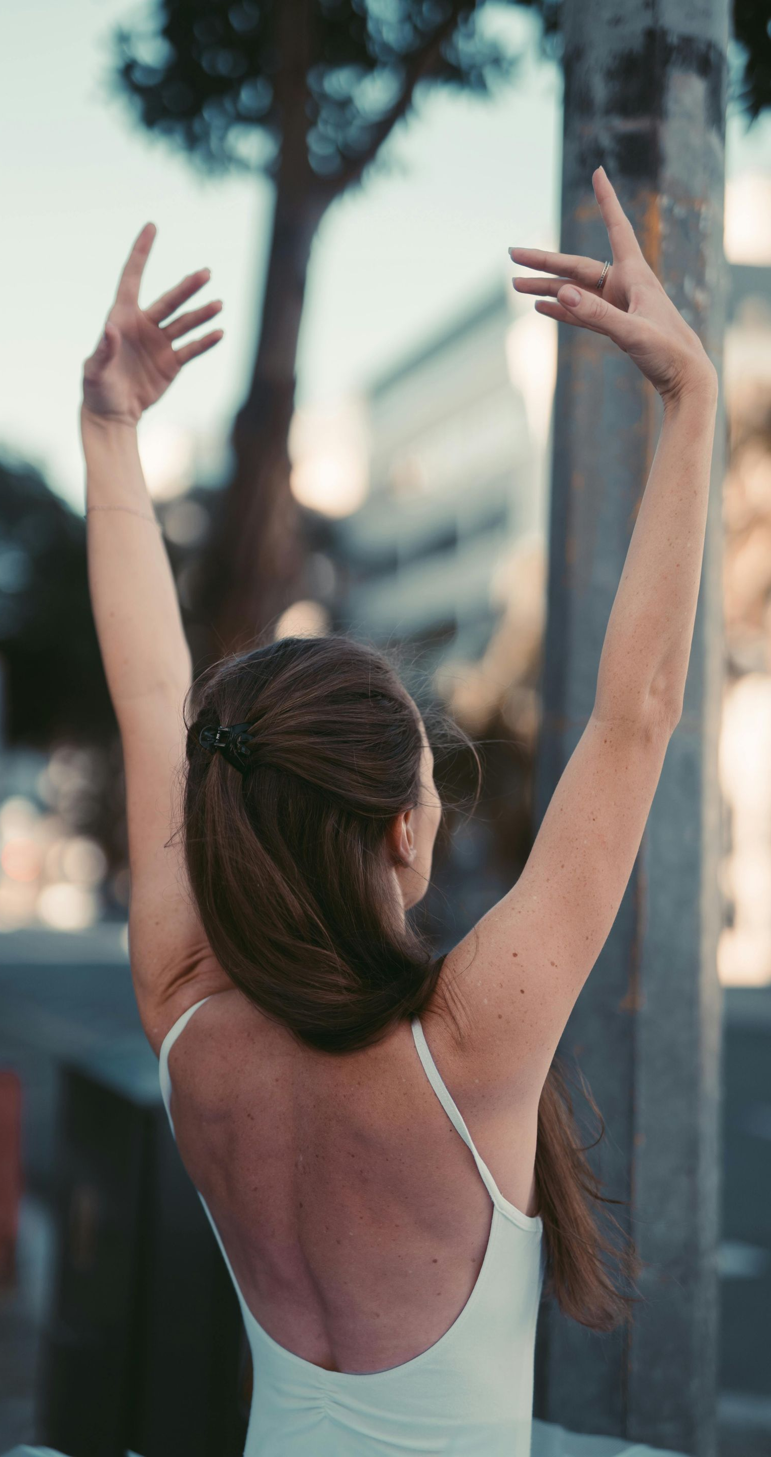 Ballerina, arms raised, back turned, outdoors. White leotard, hands gracefully posed. City background.