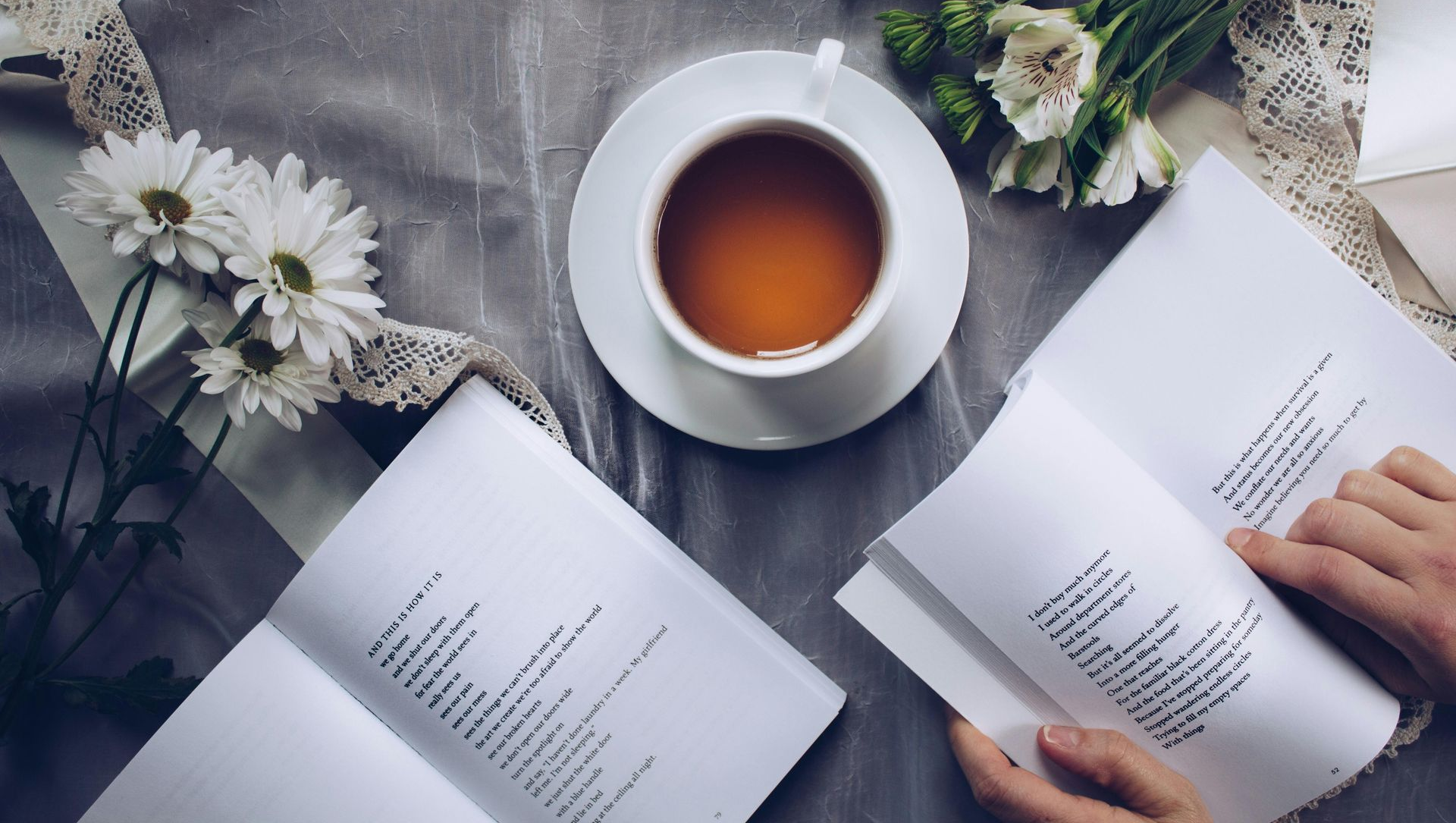 Cup of tea on saucer, open book, white flowers, lace ribbon, hands turning pages.