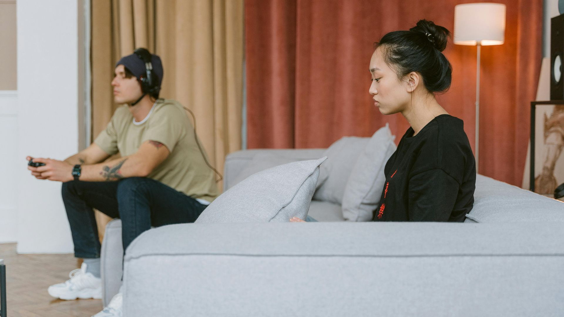 A person sits on a couch, looking away from another person playing a video game in a living room.