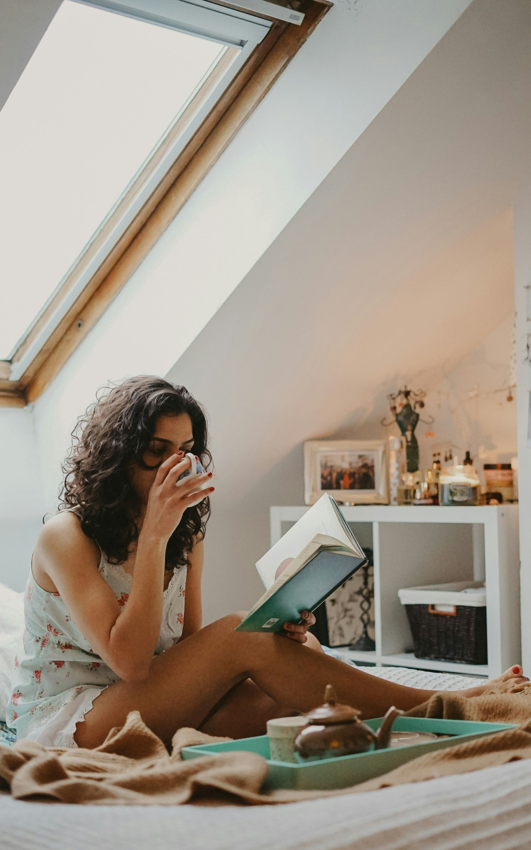 Woman in pajamas reading in bed, sipping from a glass, tray of tea nearby, skylight above.