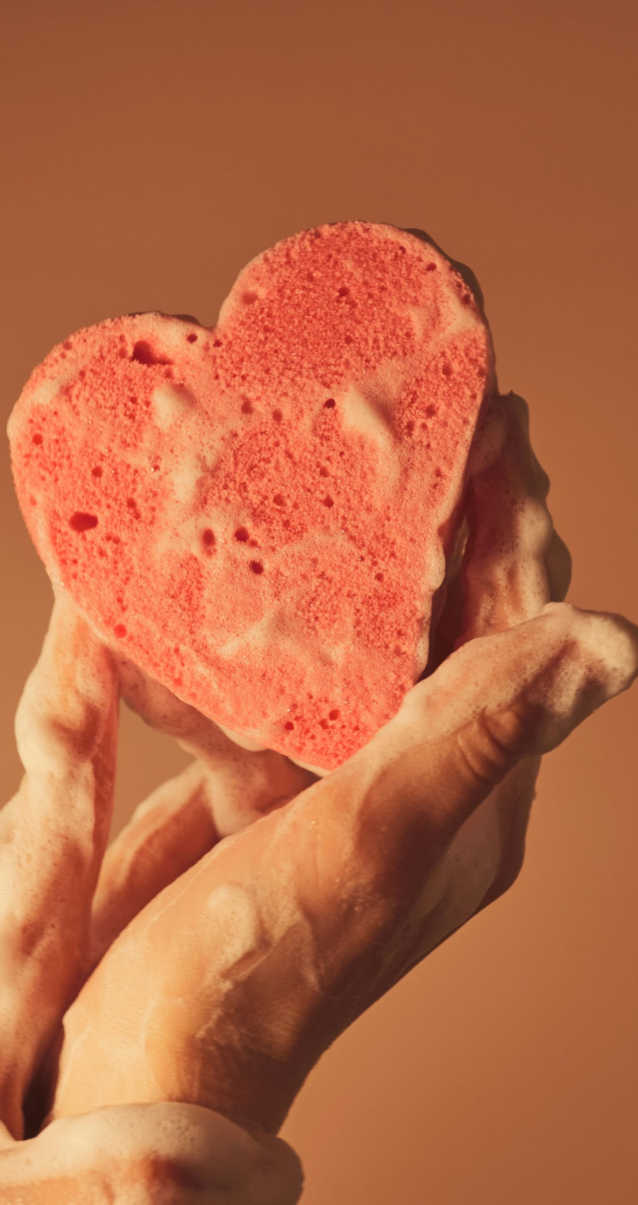 Hand holding a red, heart-shaped sponge covered in suds, against a brown background.