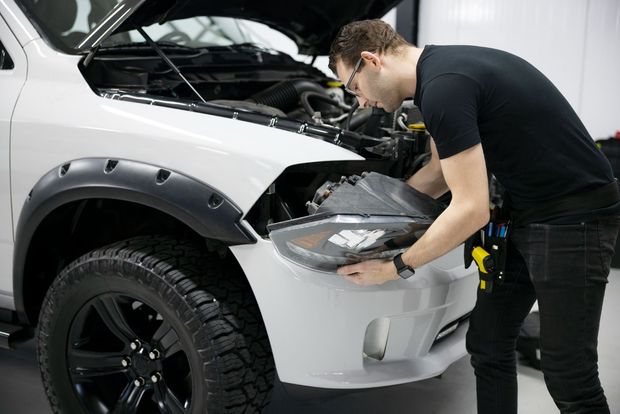 Man in black shirt removing a headlight from a white truck with open hood.
