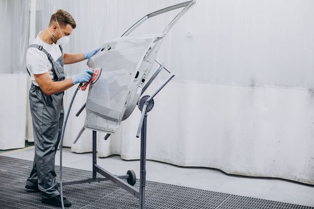Man in protective gear sanding a car door in a body shop.