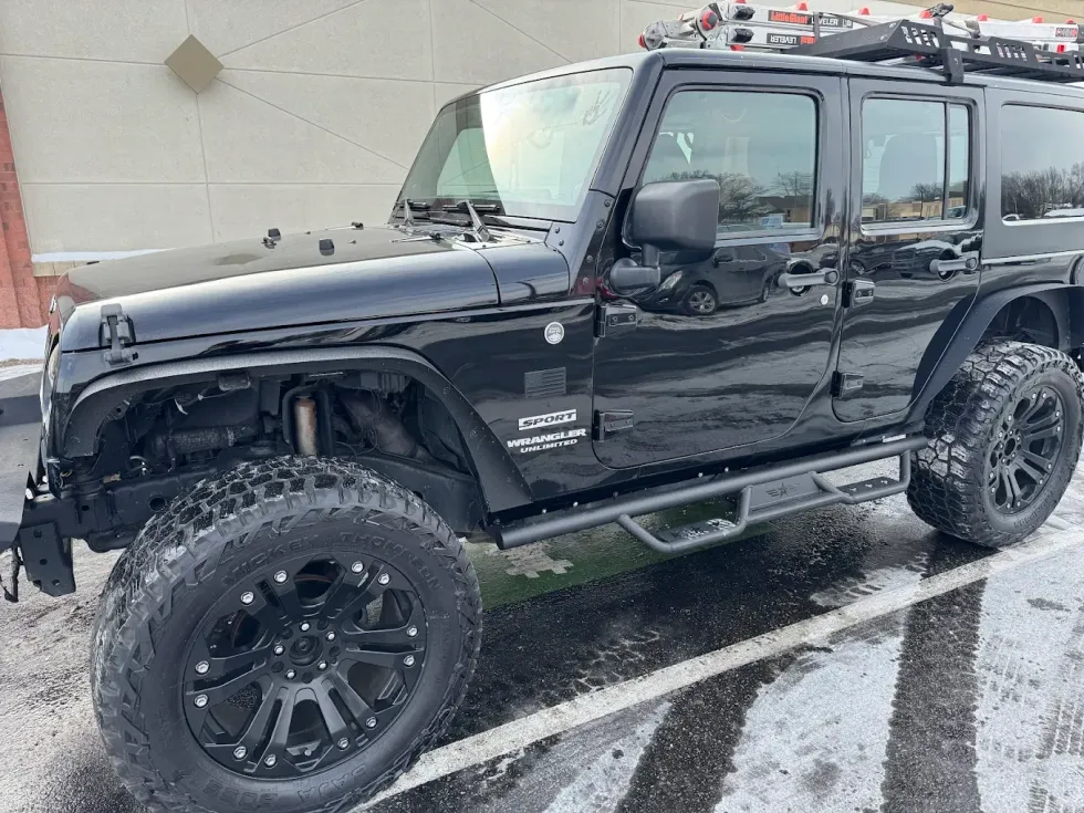 Black Jeep Wrangler parked on a snowy surface, with a roof rack and oversized tires.
