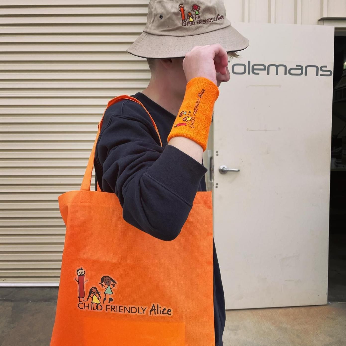 a Man Wearing a Hat and Wristbands is Holding an Orange Bag That Says Child Friendly Alice — Pinpoint In Alice Springs, NT
