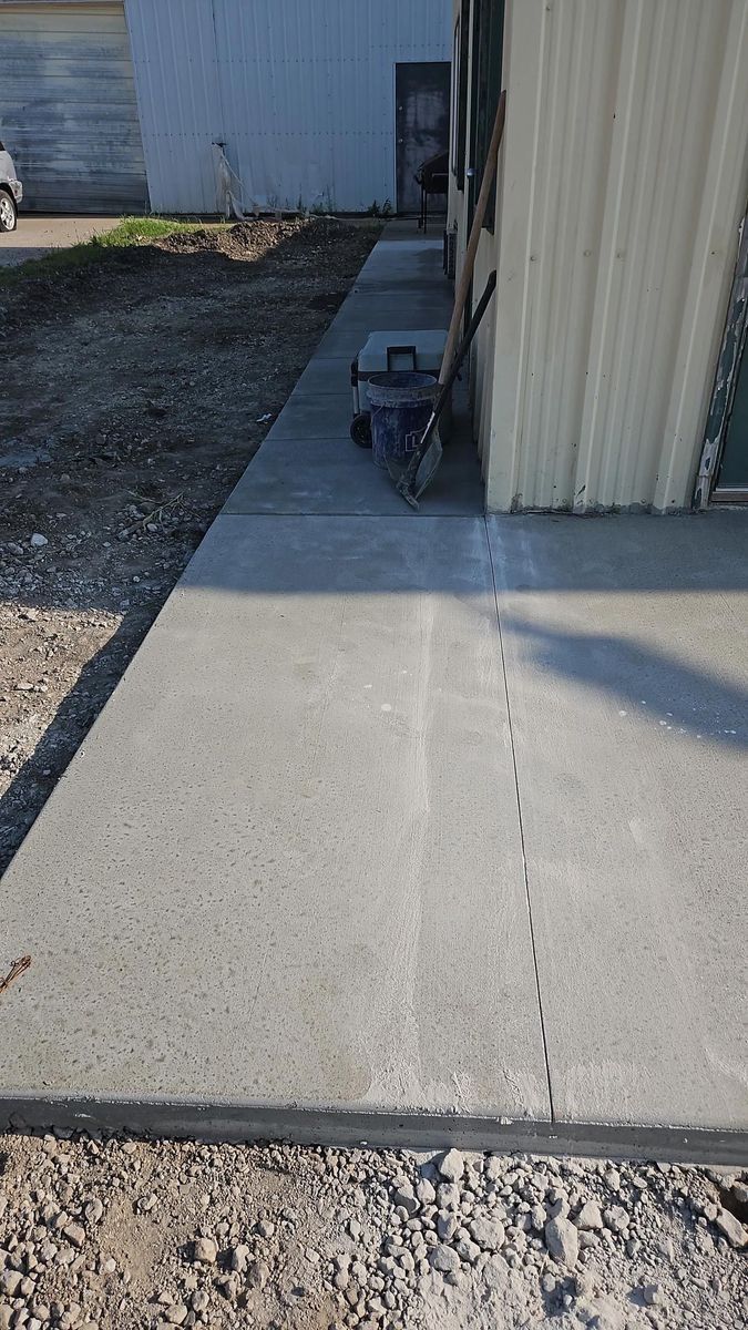 A concrete walkway leading along the side of a building, featuring a dashed control joint line down the center.