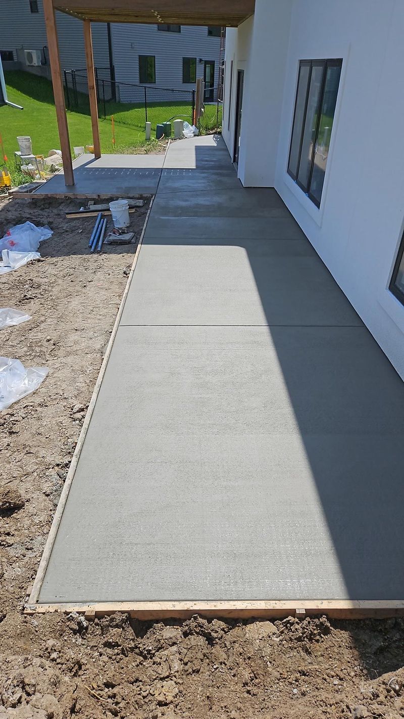 A freshly poured gray concrete walkway runs along the side of a white house on a sunny day.