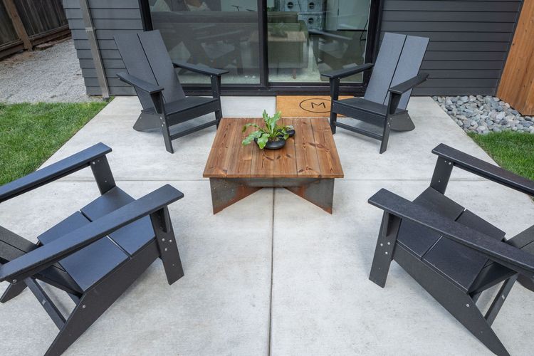 A patio set with four black Adirondack chairs surrounding a rustic wooden coffee table on a concrete slab.