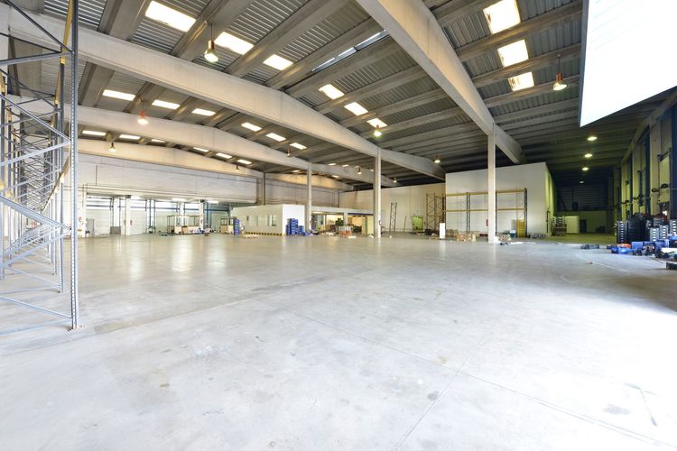 A wide-angle interior view of a large, empty industrial warehouse with high ceilings, exposed beams, and concrete floors.