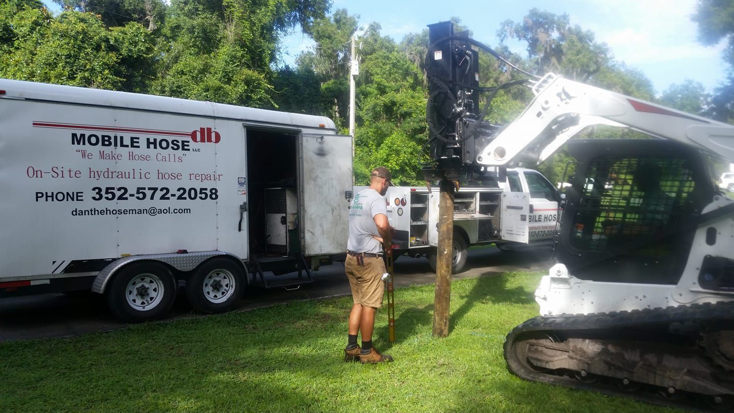Man stands near trailer and Bobcat. Utility pole being worked on. Green grass and trees in the background.