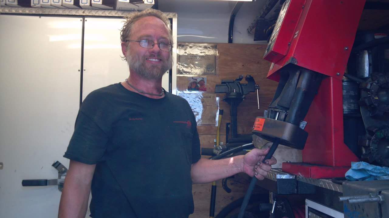 Man in a garage smiles, holding a tool near a large red machine.