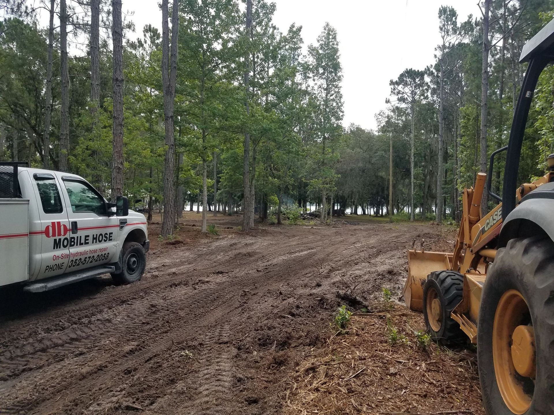Muddy construction site with a truck and backhoe surrounded by trees.