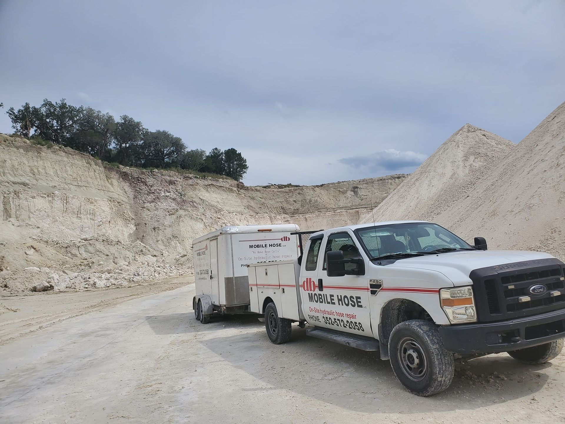 White truck with trailer on a gravel road in a quarry. Truck has company logo. Cliffs and mounds of stone in background.
