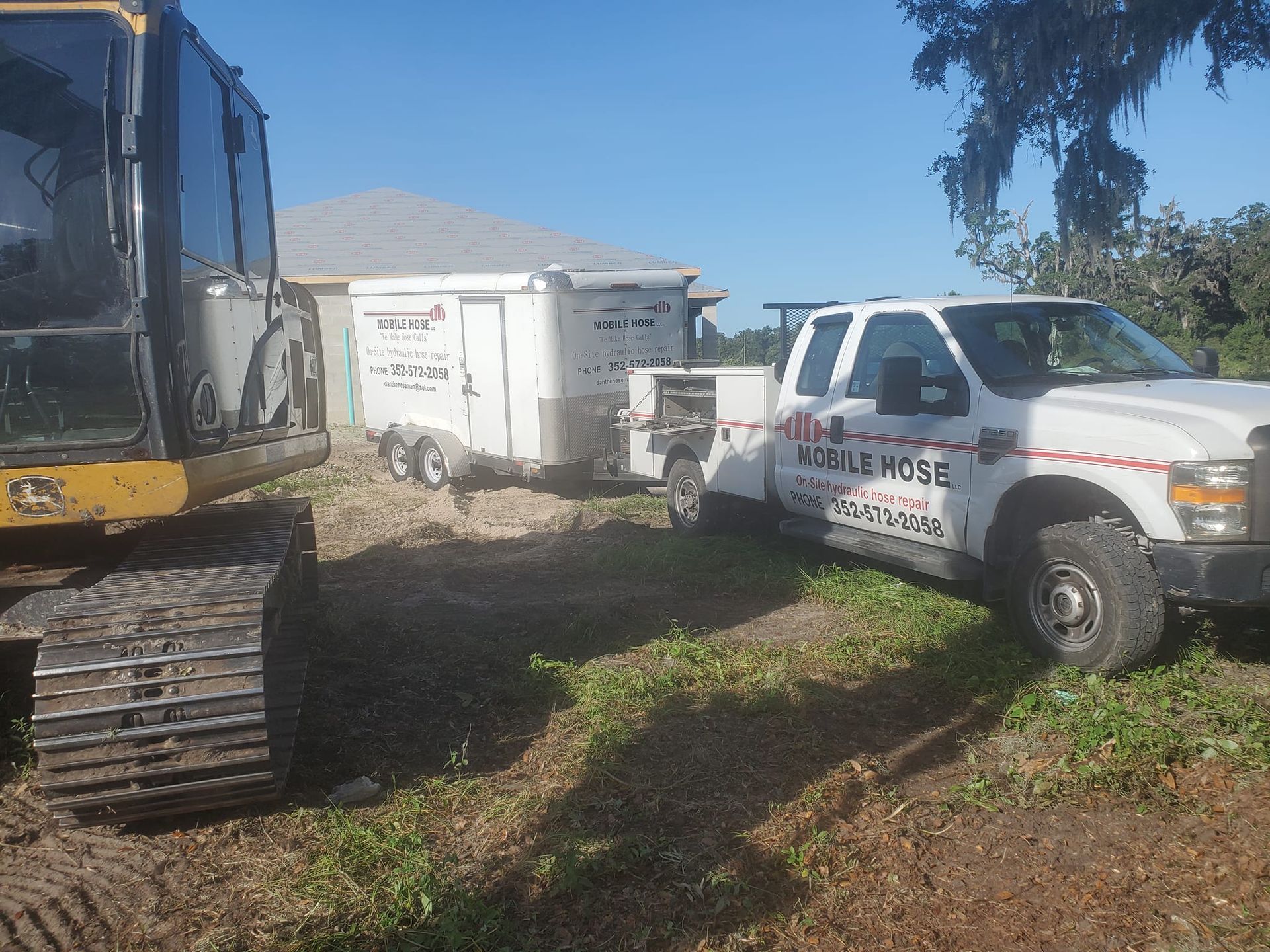 A white pickup truck, trailer, and excavator sit on a grassy area near a building on a sunny day.