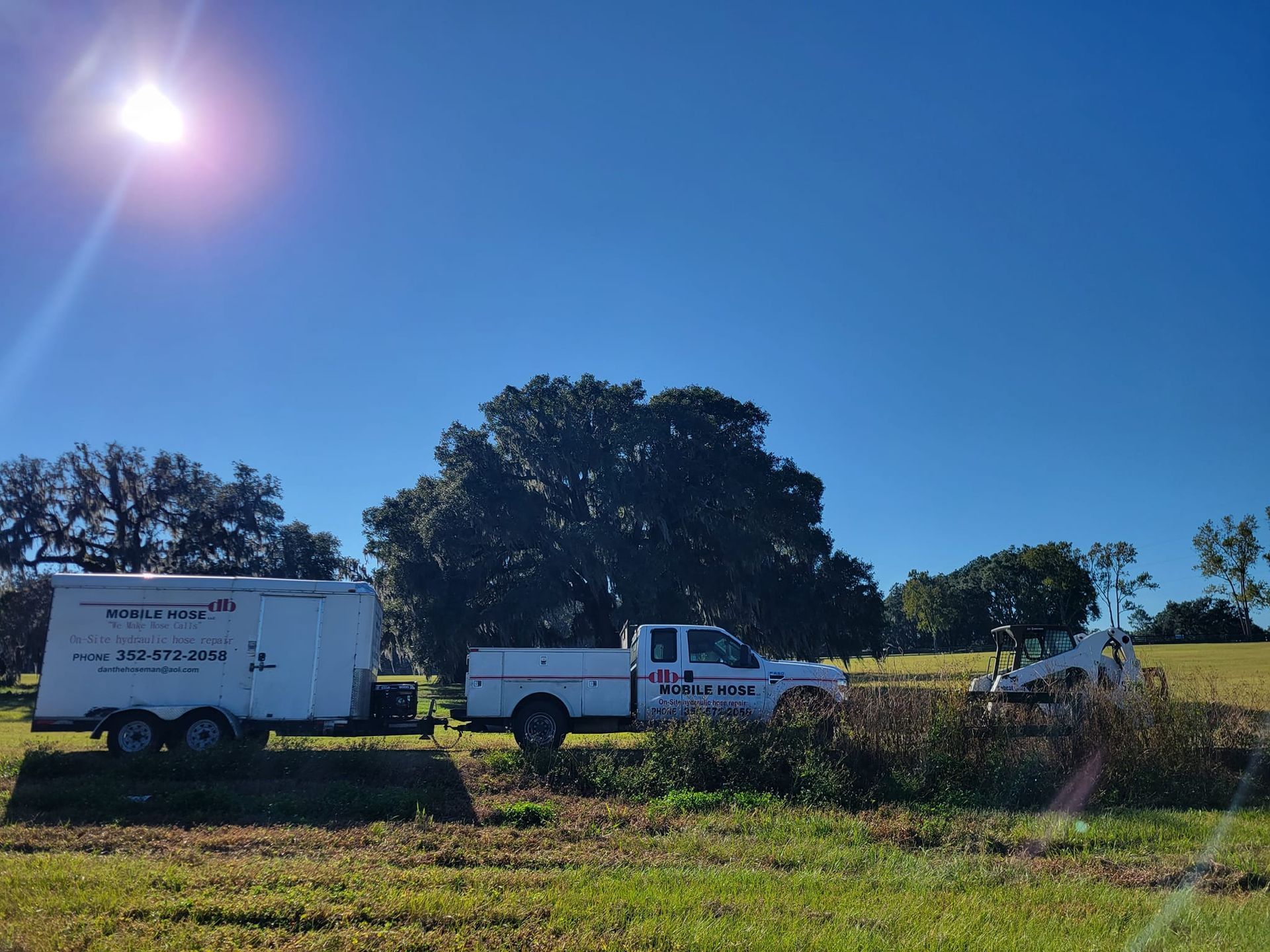 White work truck, trailer, and small bulldozer in a field under a bright blue sky.