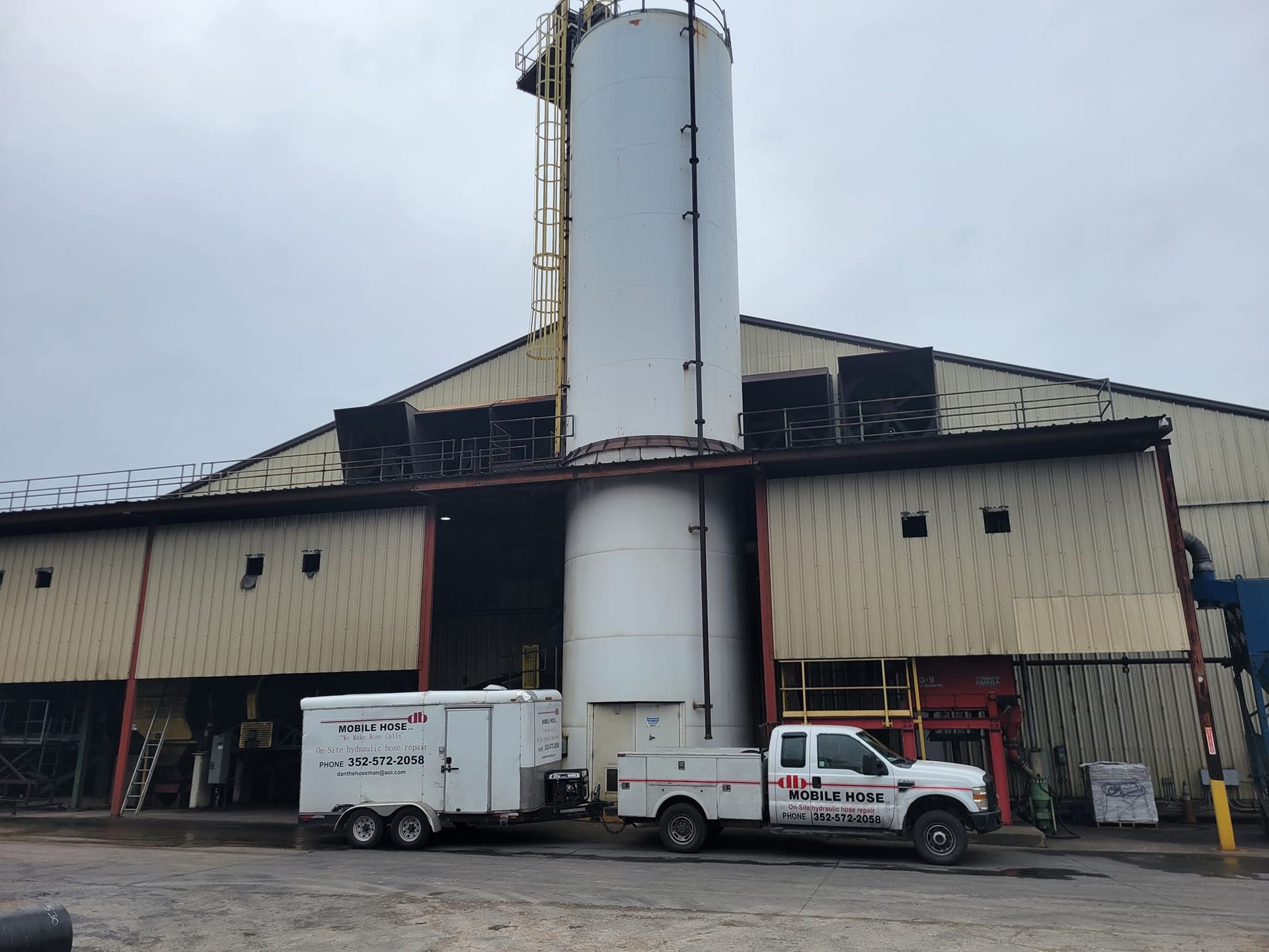 A white industrial building with a cylindrical tank, a parked truck, and a trailer on a cloudy day.