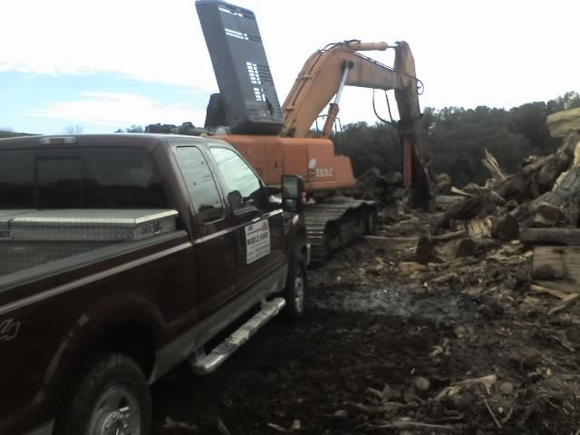 A maroon truck and orange excavator working on a pile of logs in a muddy area.