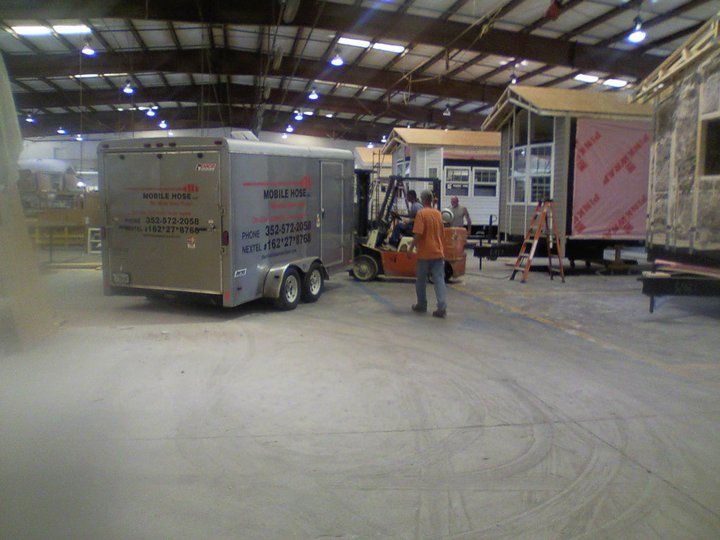Forklift moving a trailer inside a warehouse with partially built tiny homes; a person is guiding.