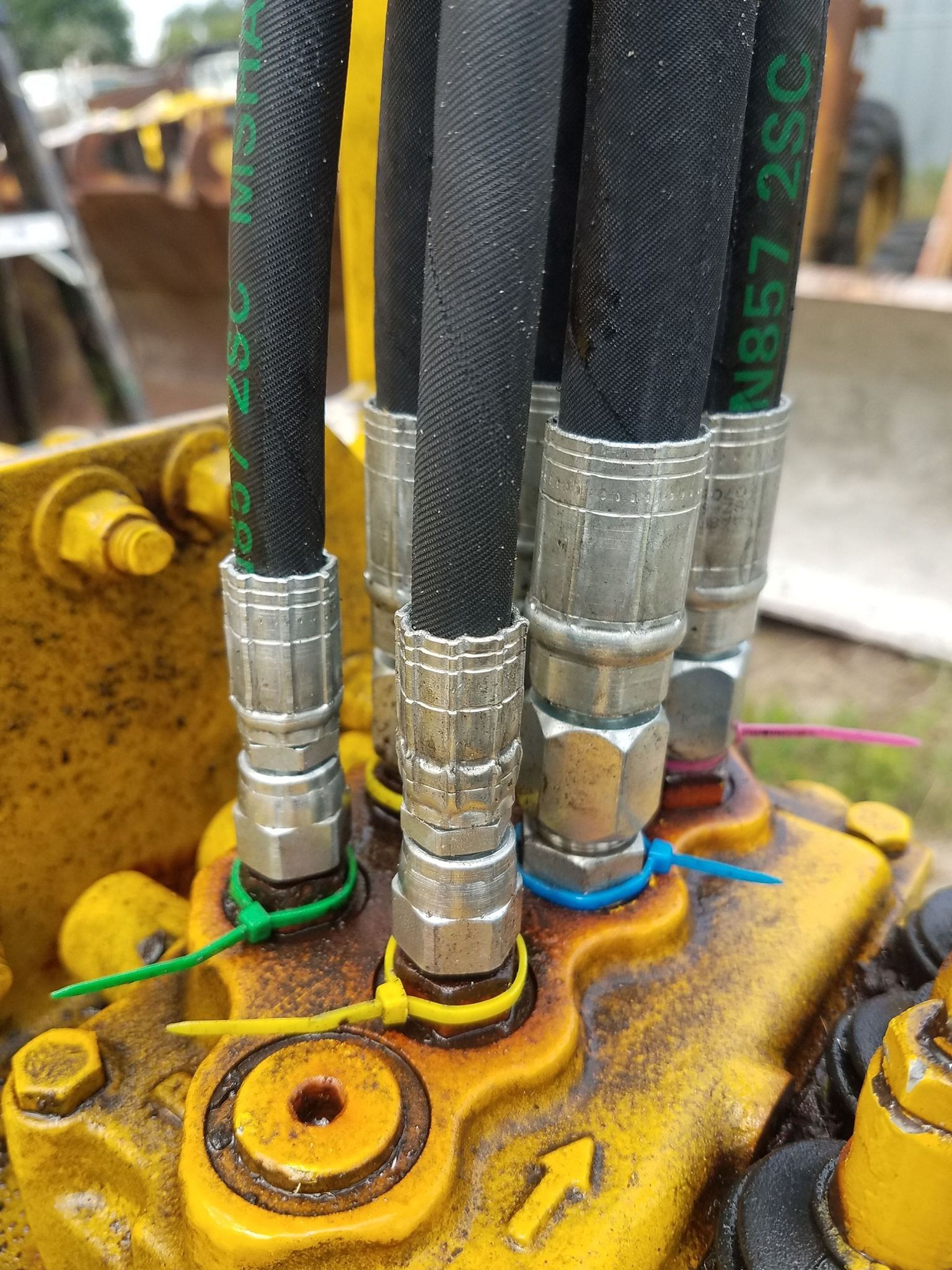 Close-up of yellow machine's hydraulic hoses connected to a control valve. Hoses are black and green, secured with zip ties.