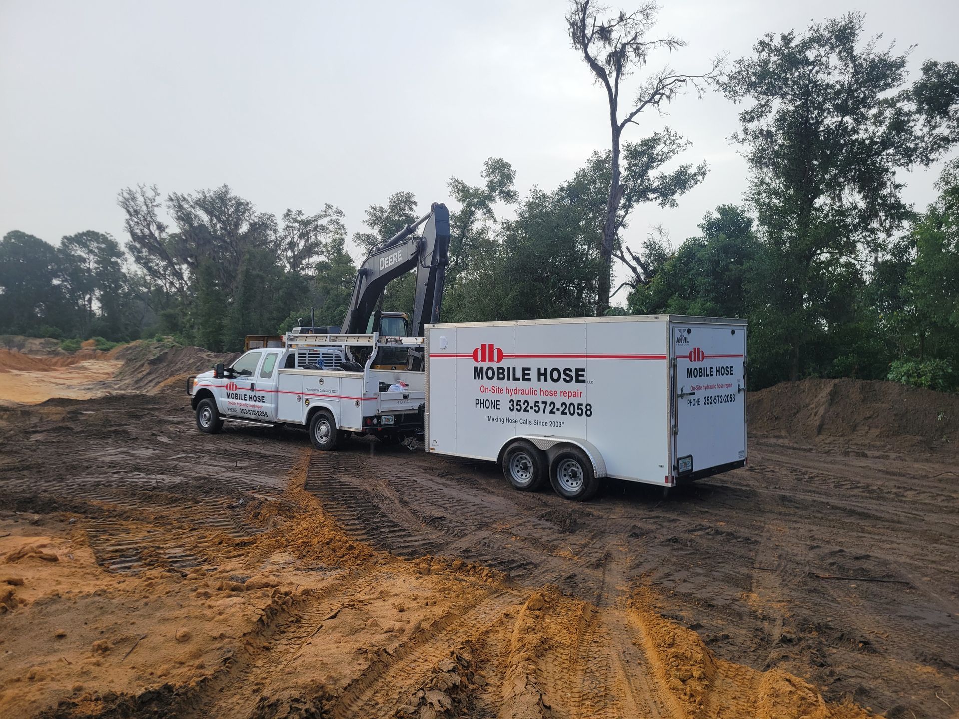 White work truck towing a trailer and excavator on a muddy construction site; overcast sky.