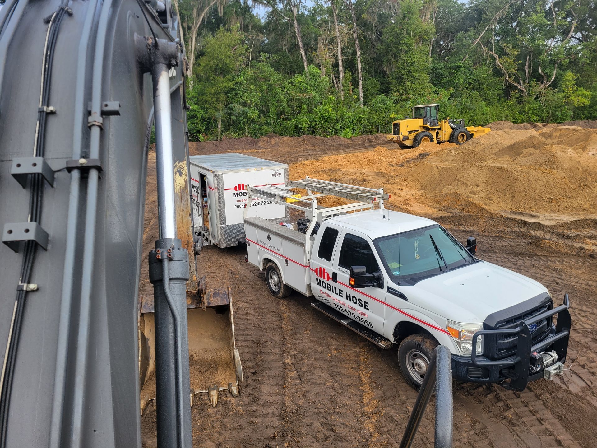 White truck towing a trailer in muddy construction site, excavator in foreground, yellow machinery in background.