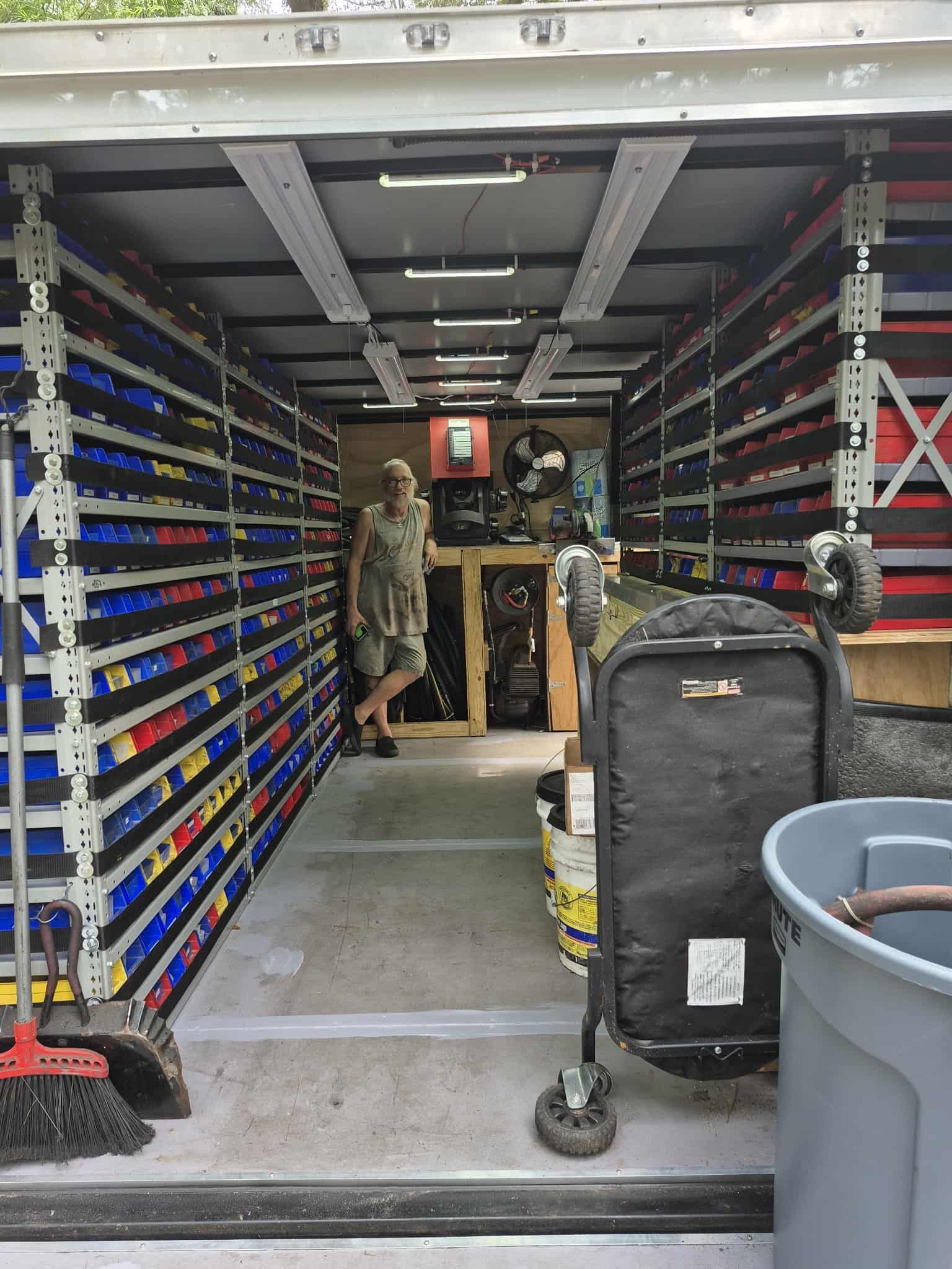 Woman stands in a storage trailer filled with shelves of parts. A cart and a trash can are in the foreground.