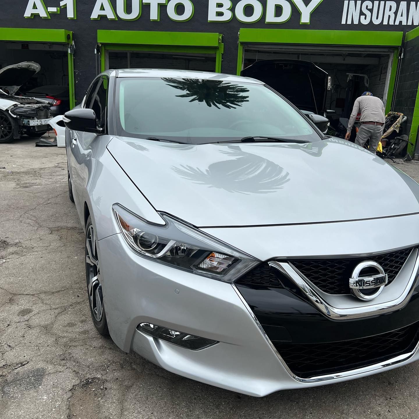 A silver nissan maxima is parked in front of an auto body shop.