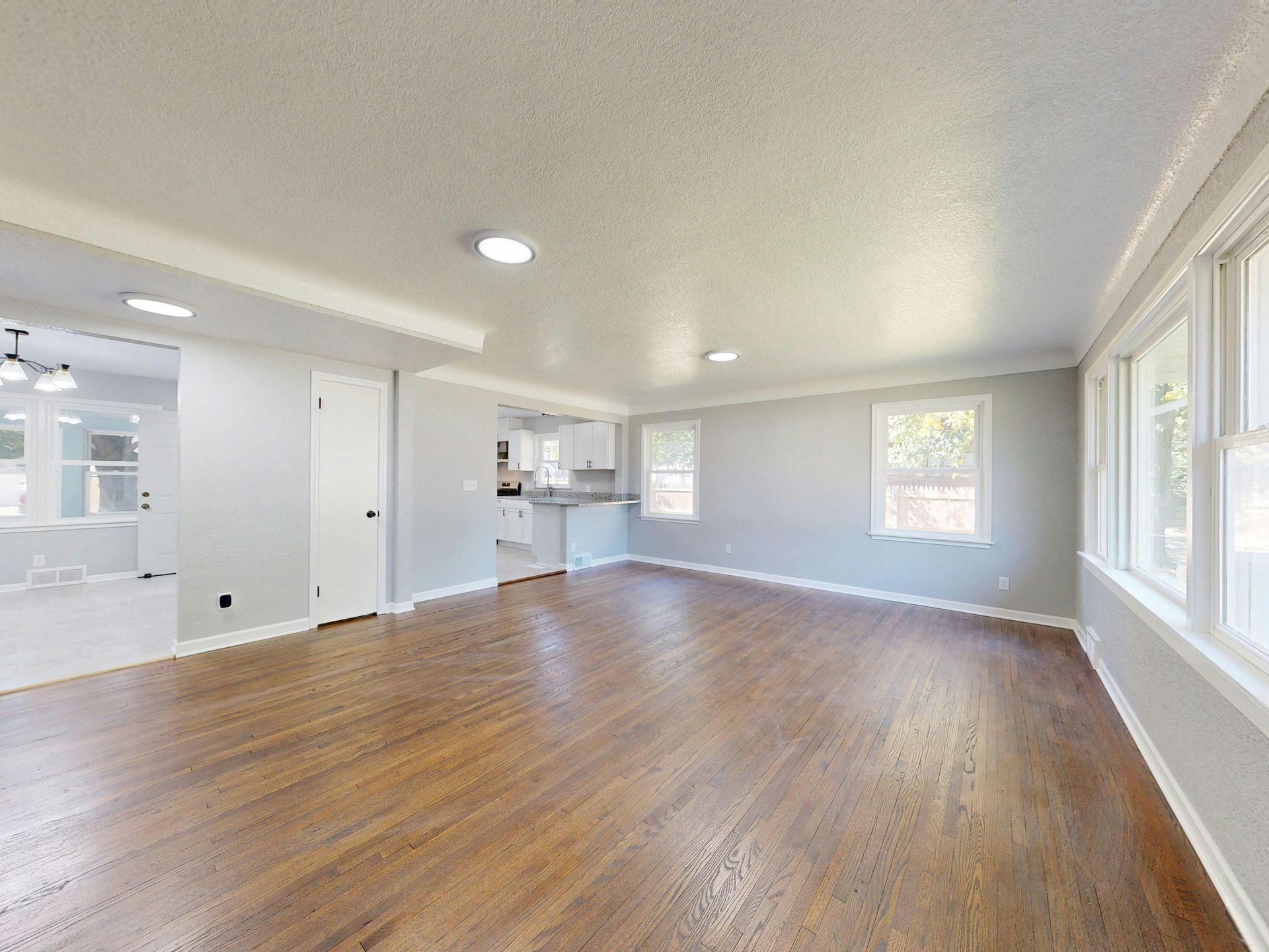 Spacious room with hardwood floors, white trim, and gray walls, leading to a kitchen.