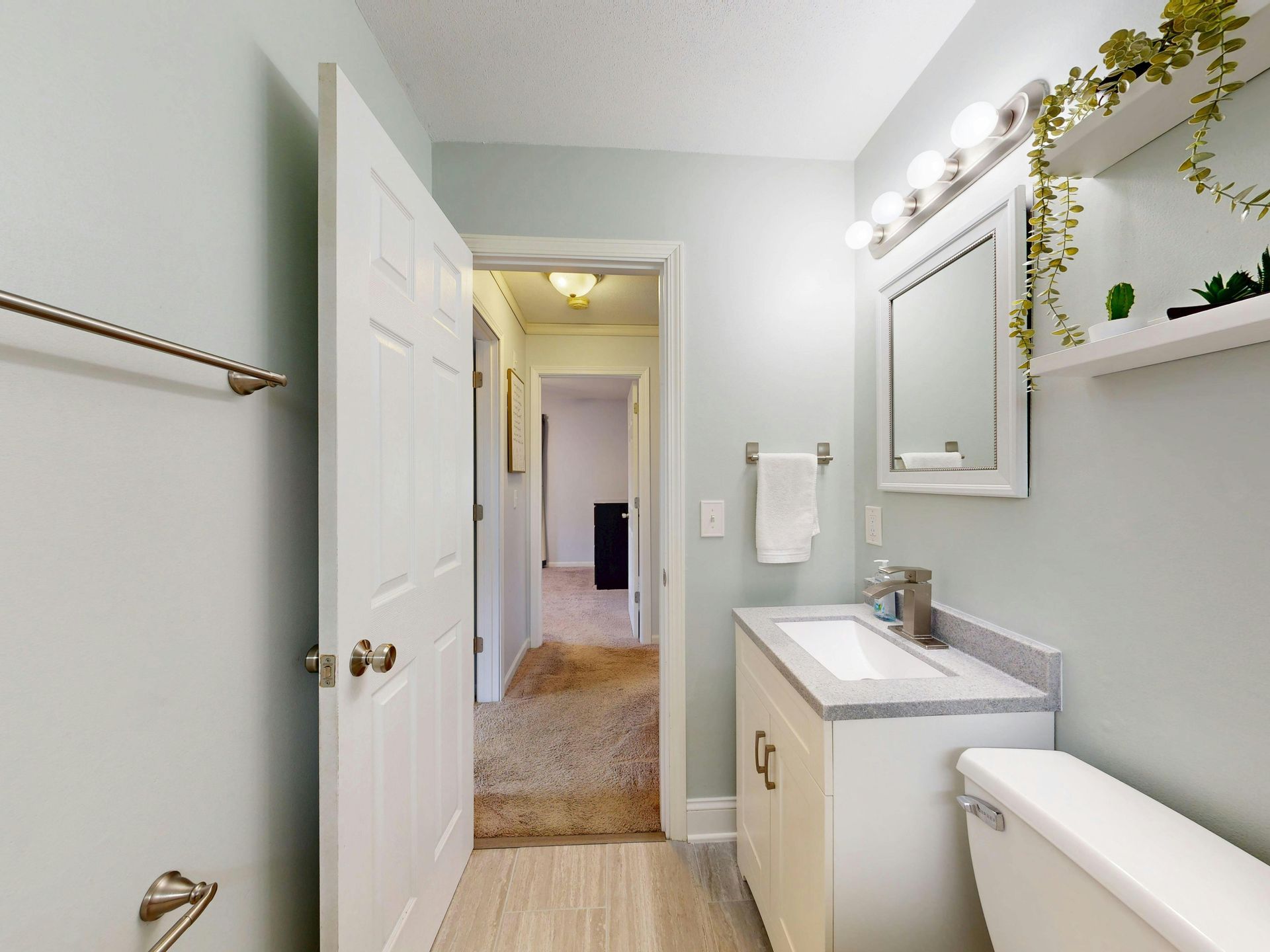 Bathroom with a white vanity, toilet, and light blue walls. Doorway leads to hallway with carpet.