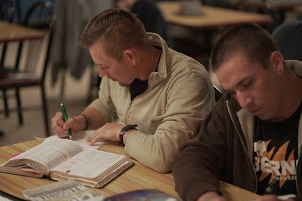 Two men are sitting at a table writing in a notebook.