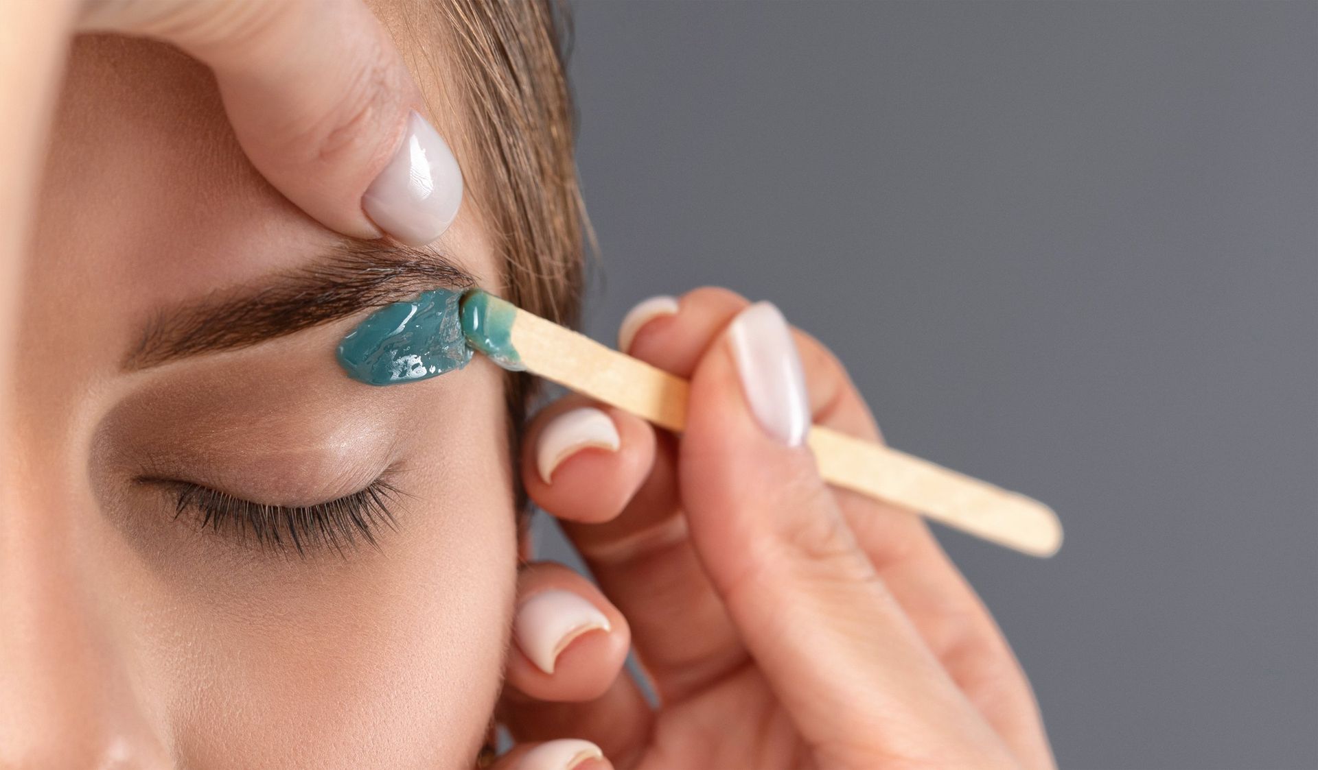 Person's eyebrow being waxed; a wooden applicator holds blue wax near the brow; gray background.