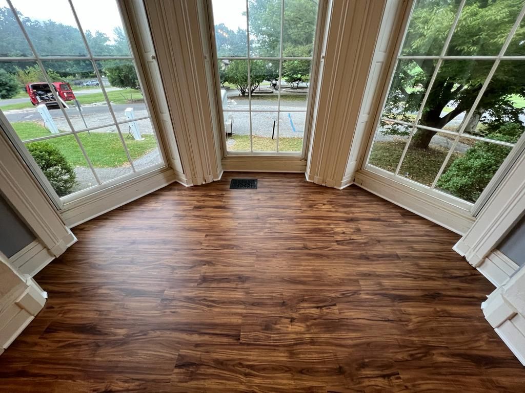 Wooden floor in a room with large windows overlooking a green yard and street.