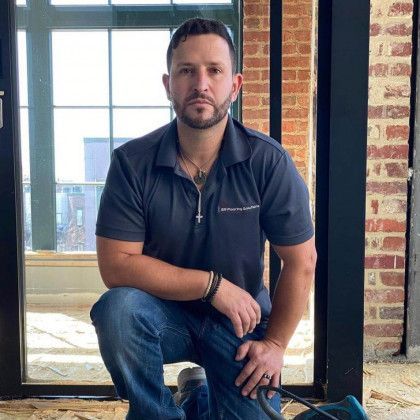 Man in navy polo and jeans kneels inside, looking at the camera. Exposed brick and window in background.