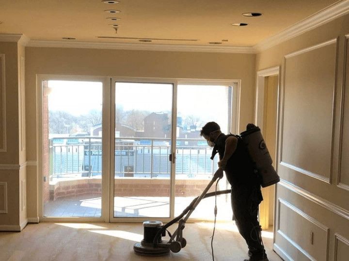 A person sanding a wooden floor in a room with large windows and balcony view.